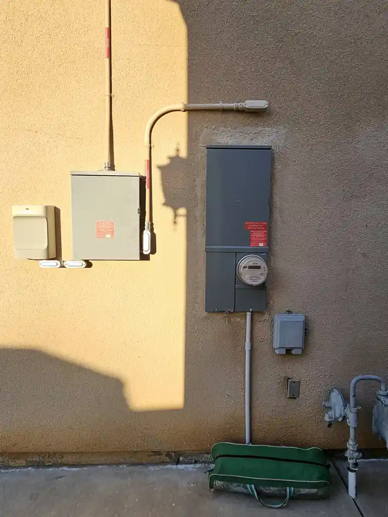 An outdoor wall with electrical panels, a utility meter, and a green duffel bag on the ground. The shadow of a plug is cast…