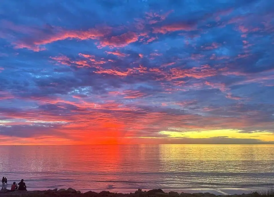 A vibrant sunset over a calm ocean with pink, orange, and blue clouds reflecting on the water; silhouettes of people are…