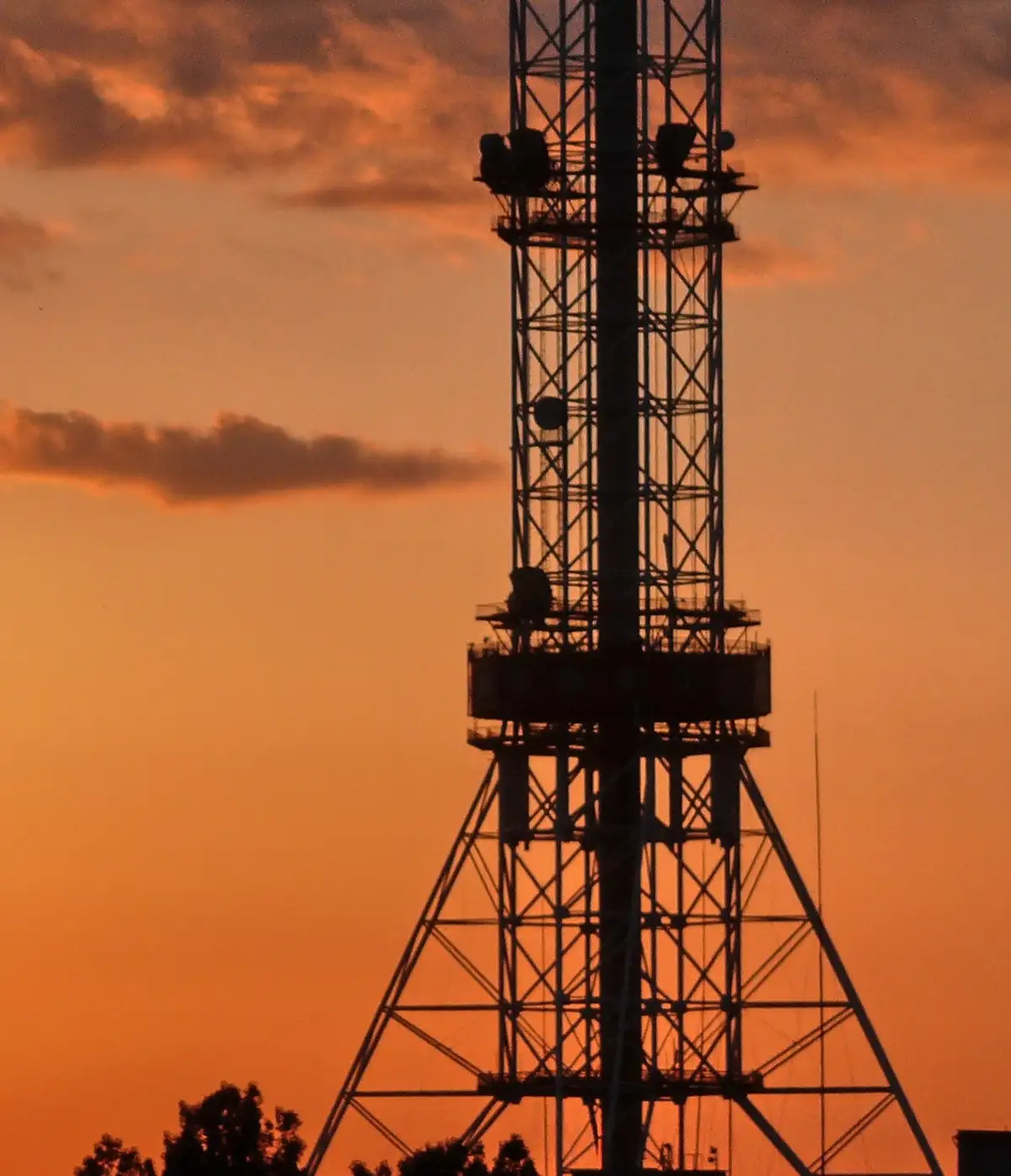 A silhouette of a tall, lattice-style communication tower against a vibrant orange sunset sky with scattered clouds. The…