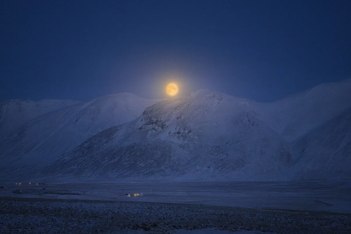 A serene, snowy mountain landscape at dusk or dawn, with a large, bright full moon rising behind a snow-capped peak. In the…