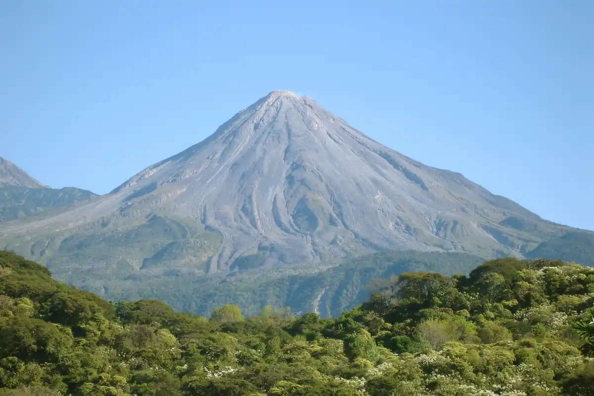 A majestic, symmetrical volcanic mountain with a gray, ash-covered peak rises against a clear blue sky, framed by lush green…