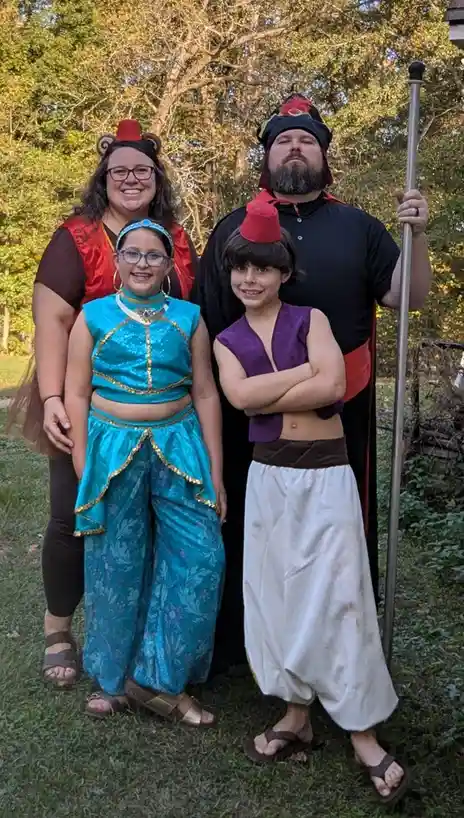 Four people pose outdoors in costumes from Aladdin: a woman as a genie with a red fez and bear ears, a girl as Jasmine in a…