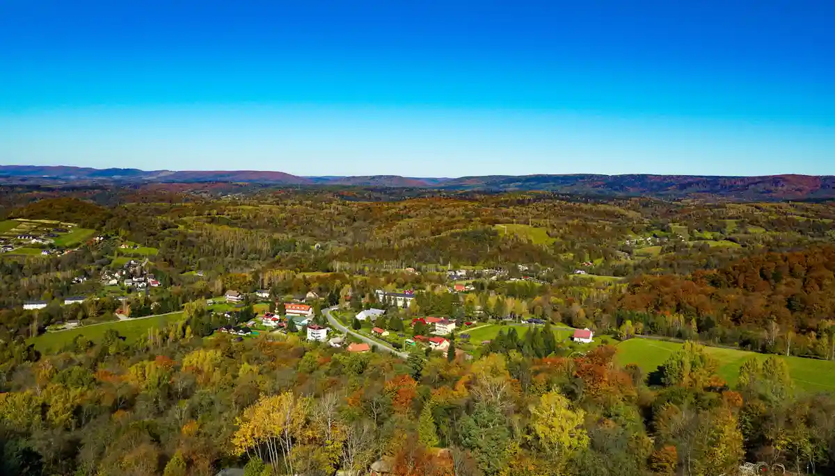 An aerial view of a picturesque village nestled in rolling hills covered in autumn-colored forests under a clear blue sky…