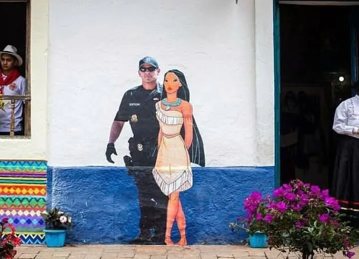 A mural on a white and blue wall depicts a real-life police officer standing beside a cartoon-style woman in traditional…