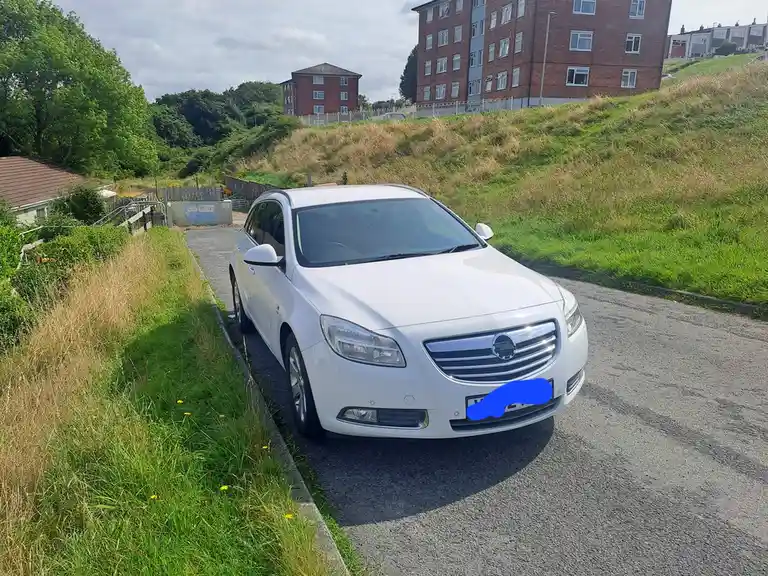 A white Opel Insignia estate car is parked on a narrow residential road next to a grassy verge, with brick apartment…