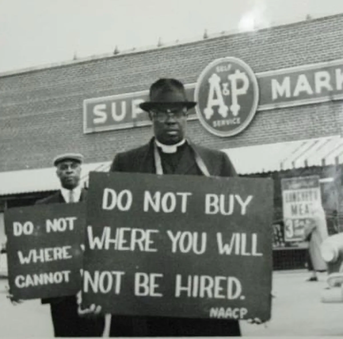 A black-and-white historical photo shows two Black men in suits and hats holding protest signs outside an A&P supermarket…