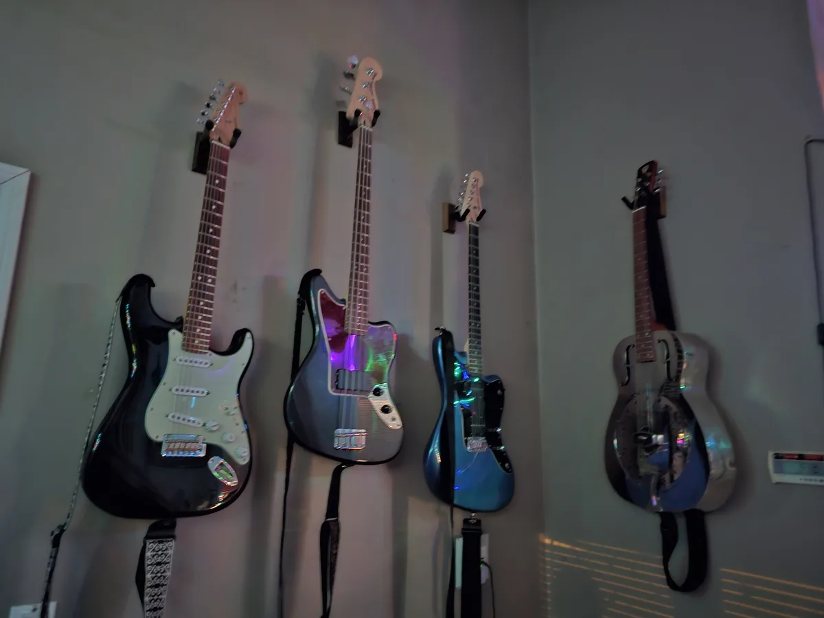 Four electric guitars and one resonator guitar are mounted vertically on a plain wall, illuminated by colorful ambient…