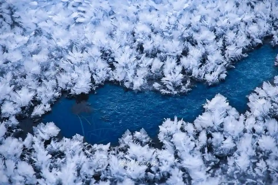 An aerial view of a vibrant blue river winding through a landscape covered in intricate, feathery frost patterns. The image…