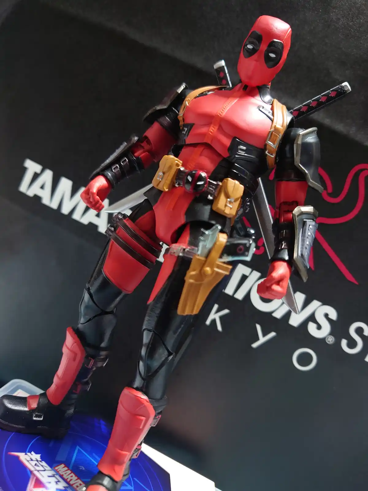 A red and black Deadpool action figure stands in a dynamic pose, equipped with multiple weapons including swords, knives…