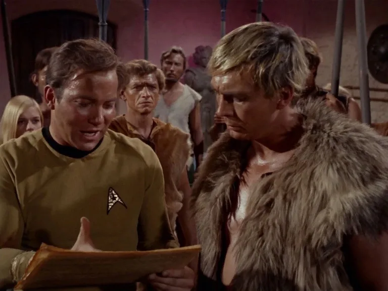 A man in a Star Trek uniform holds a scroll and speaks to a man in a fur vest, who looks downcast. Other people stand in the…