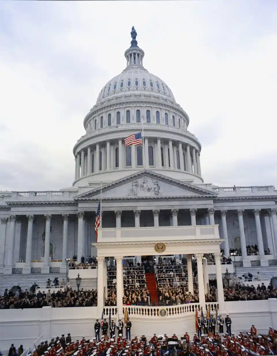 A historical photograph of the U.S. Capitol building with crowds gathered on the steps and a military band in front. The…
