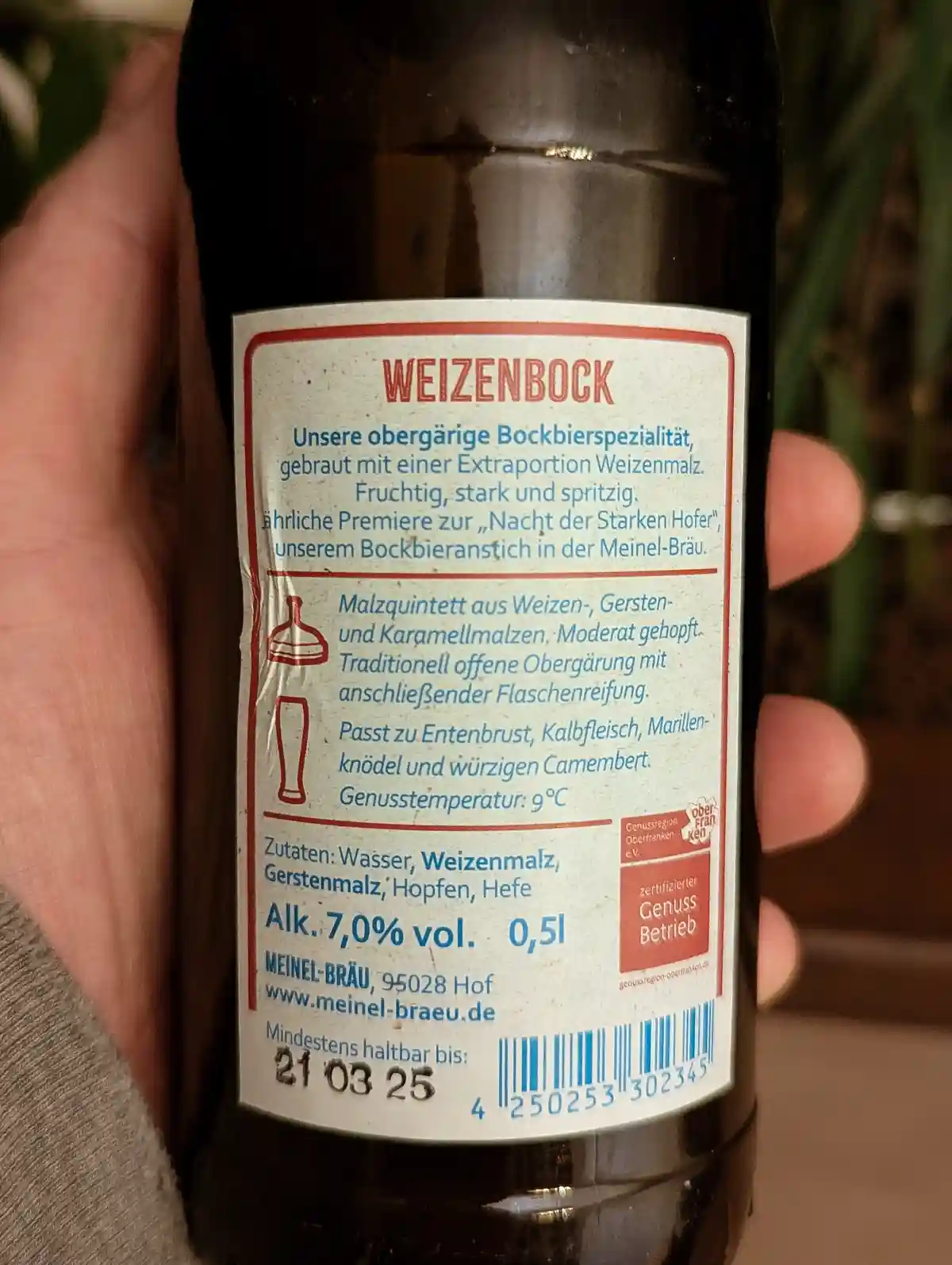 A close-up photo shows a hand holding a dark glass bottle of Weizenbock beer, with its label clearly visible. The label is…