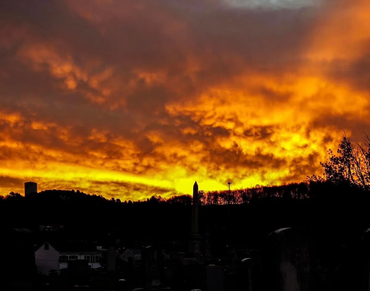 A dramatic sunset with fiery orange and yellow clouds over a silhouette of a cemetery with tombstones and a distant obelisk…