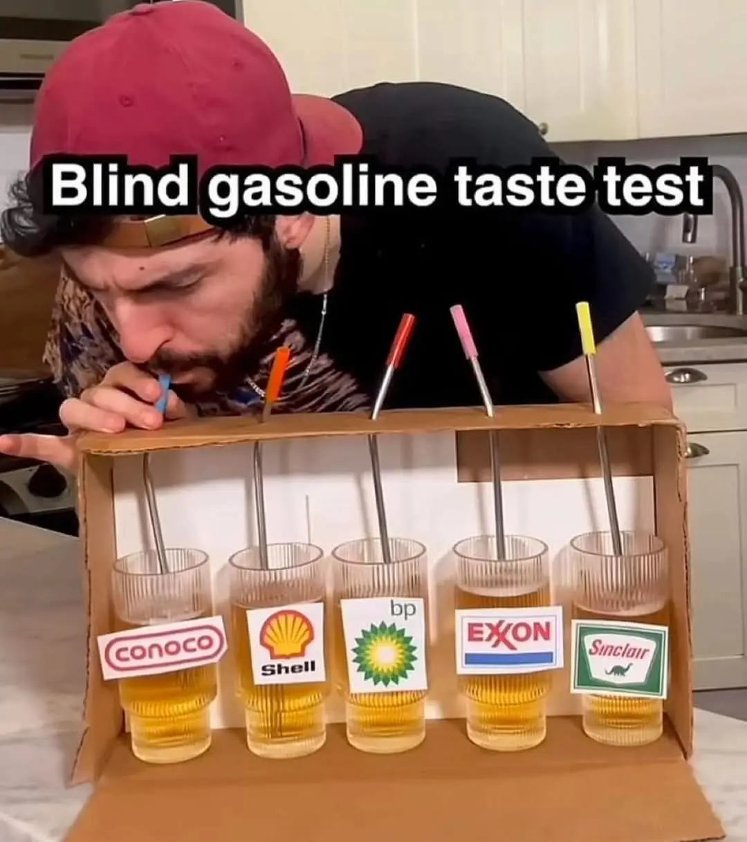 A man in a red cap leans over a cardboard tray holding five glasses filled with a yellow liquid, each labeled with a…