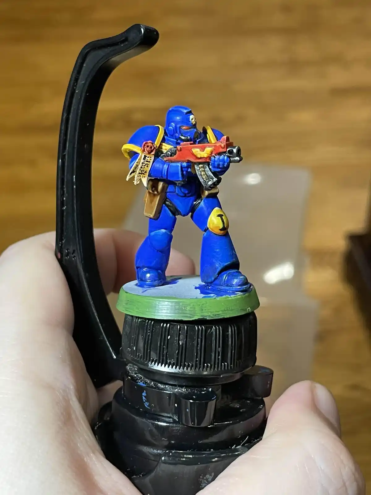 A hand holds a black plastic bottle cap with a miniature blue Space Marine figurine painted in blue, yellow, and red…