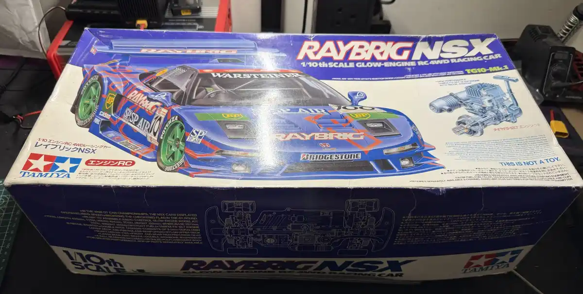 A close-up photo shows the box of a Tamiya 1/10th scale glow-engine RC 4WD racing car model, specifically the Raybrig NSX…