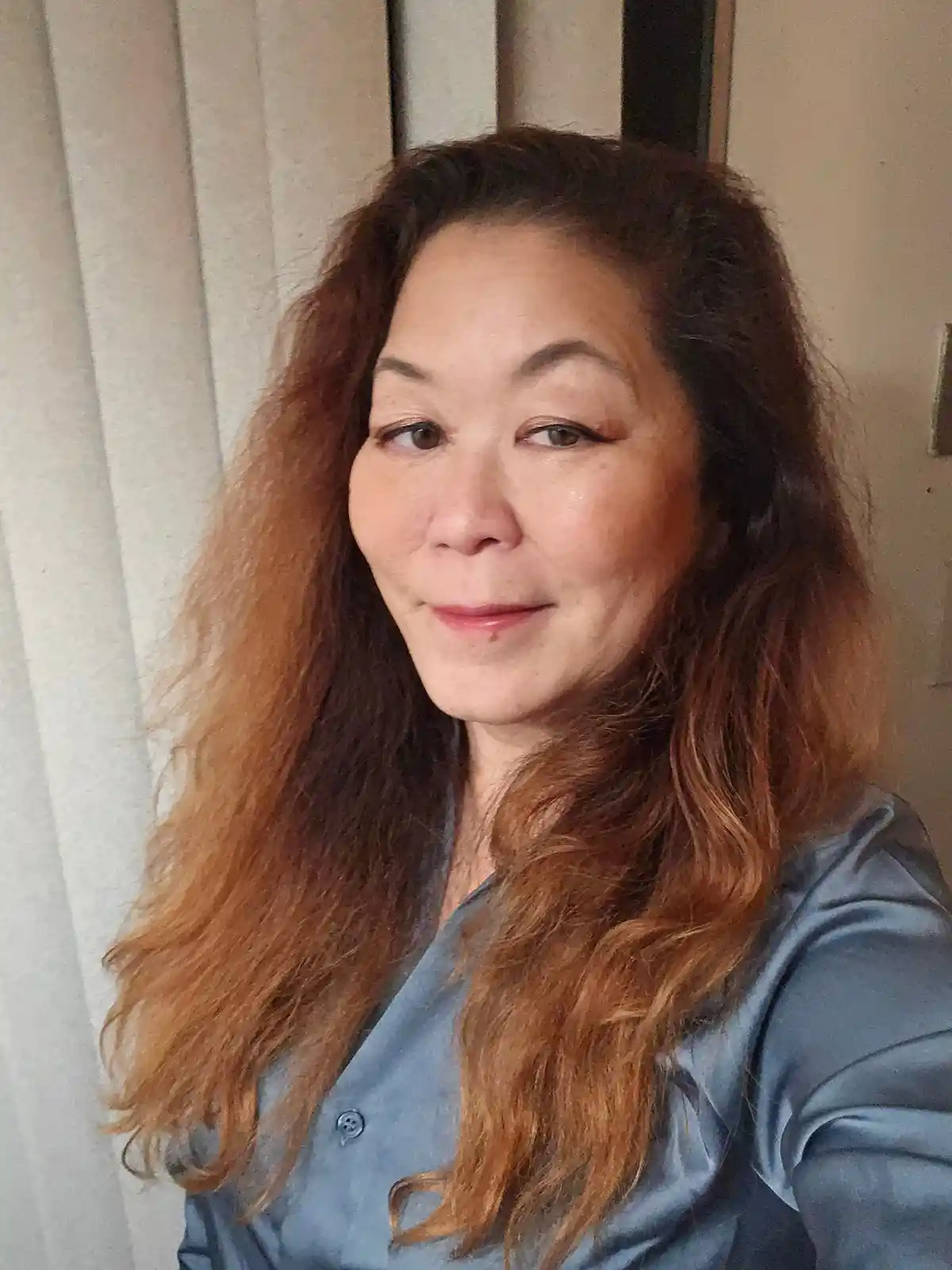 A woman with long, wavy, reddish-brown hair and a subtle smile looks at the camera while wearing a blue satin shirt, with…