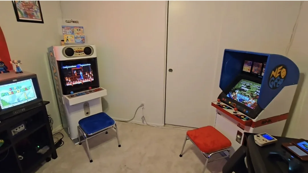 A room with three arcade machines: a white 'Caton' machine on the left, a blue 'Neo Geo' machine on the right, and a TV…