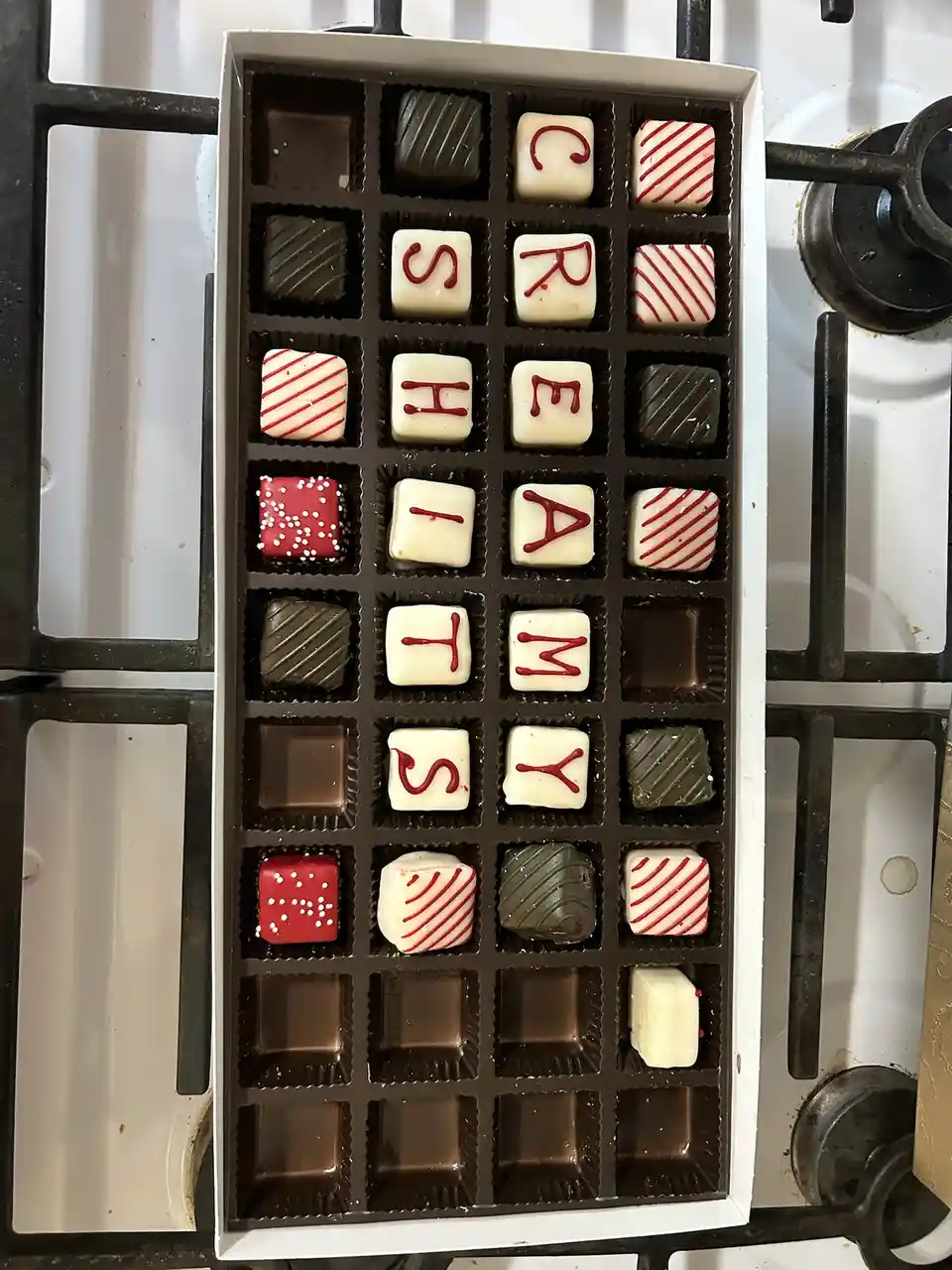 A box of assorted chocolates sits on a stovetop, with some pieces decorated with red icing letters spelling out 'CRUSH MY…