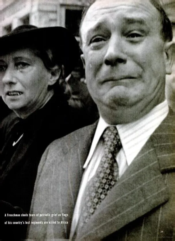 A black-and-white historical photograph shows a man in a suit and tie with a tearful expression, standing beside a woman in…