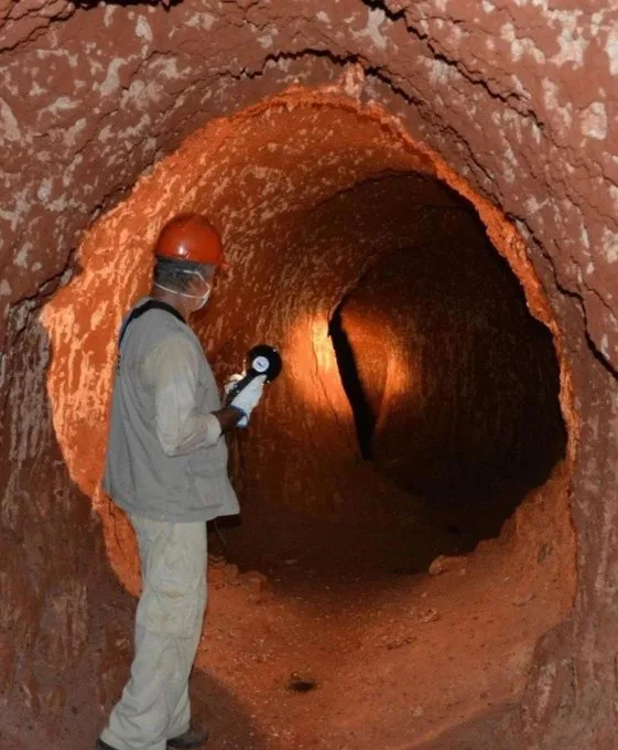 A person wearing a hard hat, face mask, and gloves stands inside a narrow, reddish-brown tunnel, holding a flashlight that…