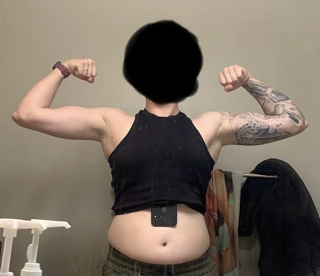 A person with a tattooed right arm and a black crop top is flexing both biceps in front of a mirror, with their face…
