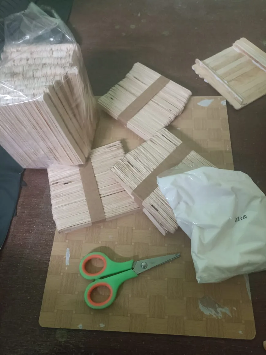 A cluttered work surface holds multiple large stacks of wooden craft sticks, some bundled with brown tape, a pair of…