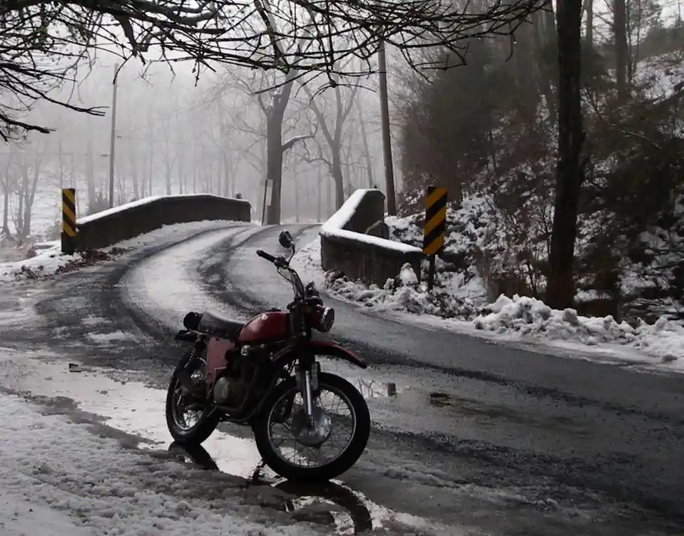 A red motorcycle is parked on a snowy, icy road next to a bridge in a foggy, wooded area. The road curves ahead, flanked by…