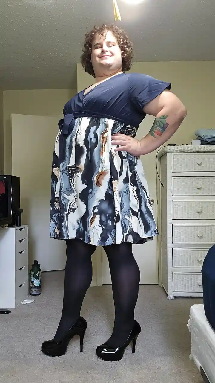 A person with curly hair and a tattoo on their left arm stands in a bedroom wearing a blue top, a patterned dress with…
