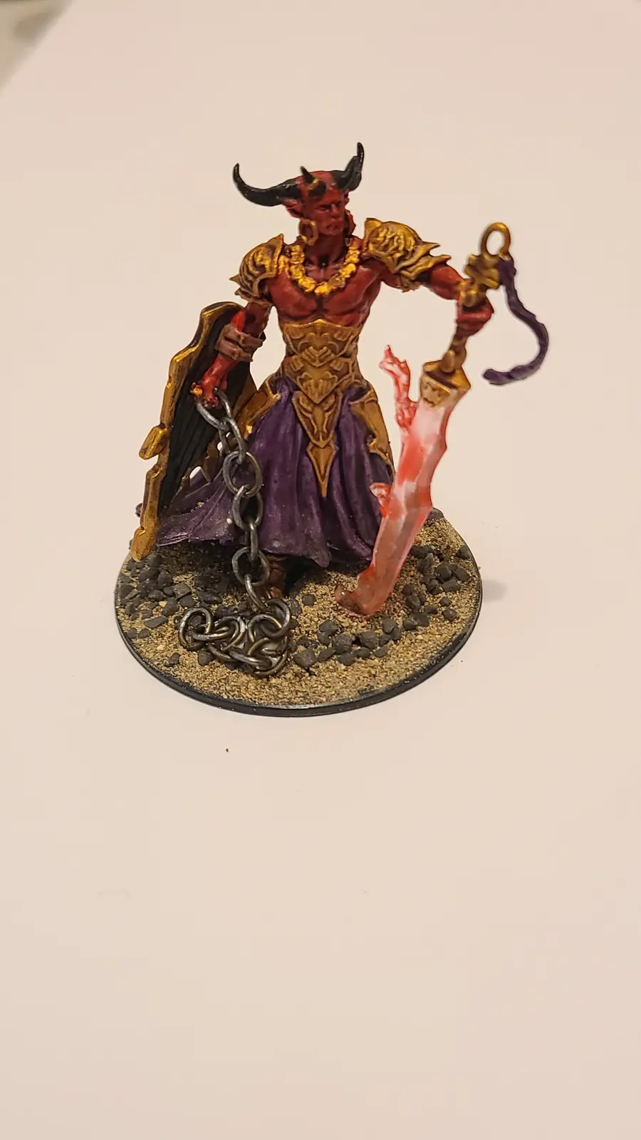 A painted miniature of a red-skinned demon with black horns, wearing gold armor and a purple skirt, holding a sword and a…