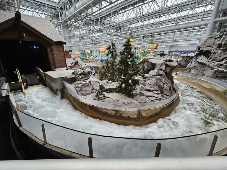 A wide-angle shot of a large, indoor water ride with turbulent, foamy water flowing around a central rocky island with a…