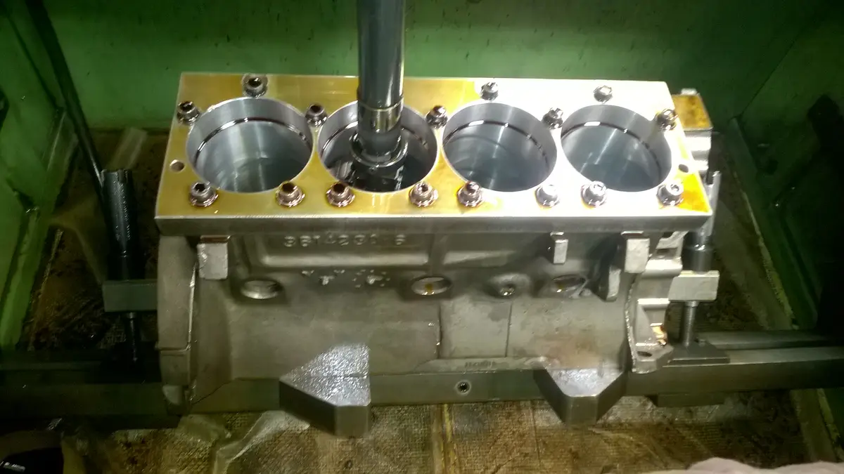 A close-up photo of a shiny, freshly machined engine block with four cylinders and a central piston rod, secured in a…