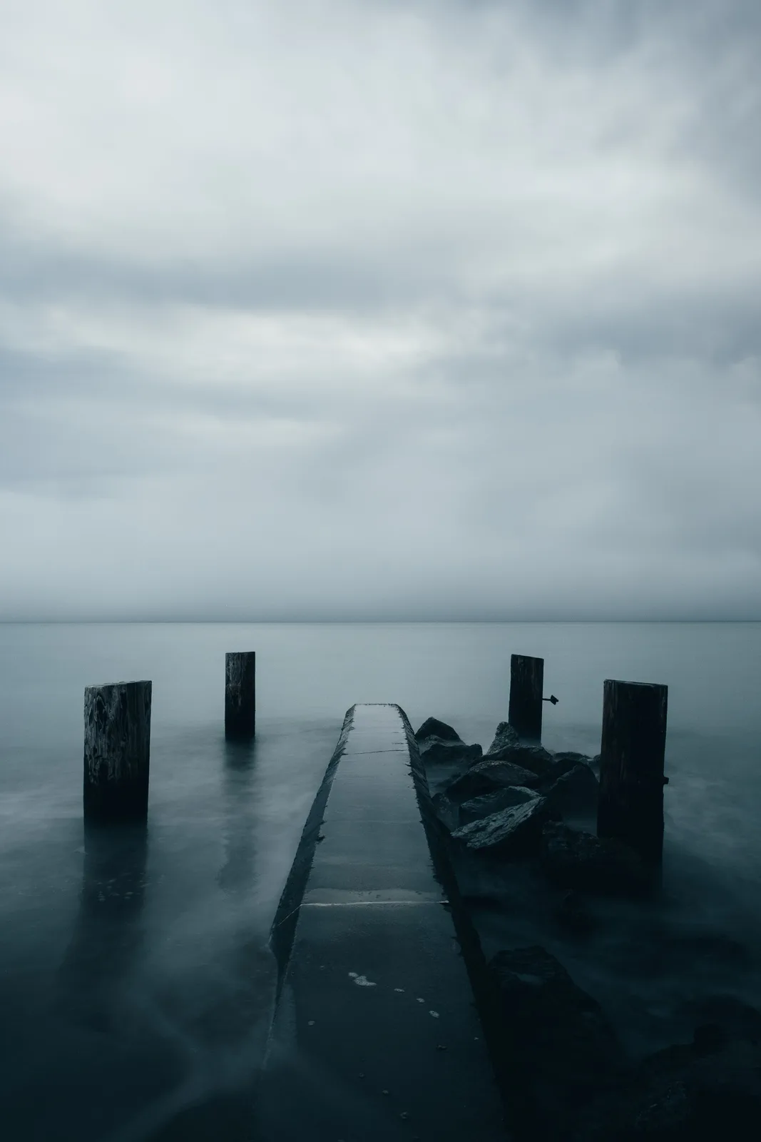 A moody, long-exposure photograph shows a narrow concrete pier extending into calm, misty water under an overcast sky…