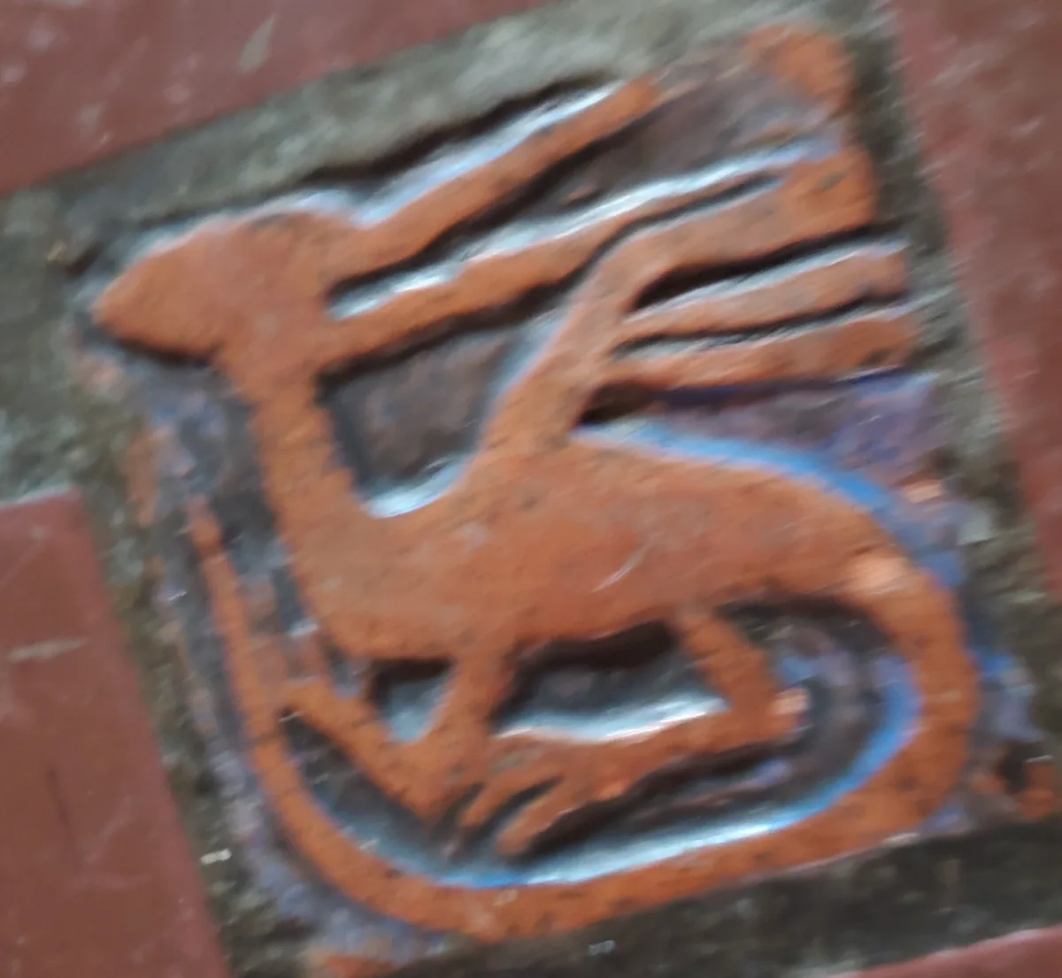 A blurry, close-up photo of a reddish-brown, stylized horse-like figure carved into a dark stone or metal plaque, with blue…