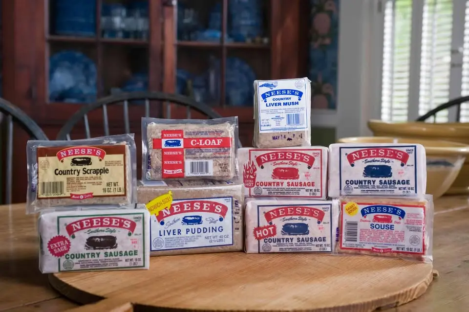 A wooden table displays a pyramid of various packaged Nee's Southern Style meat products including scrapple, liver pudding…