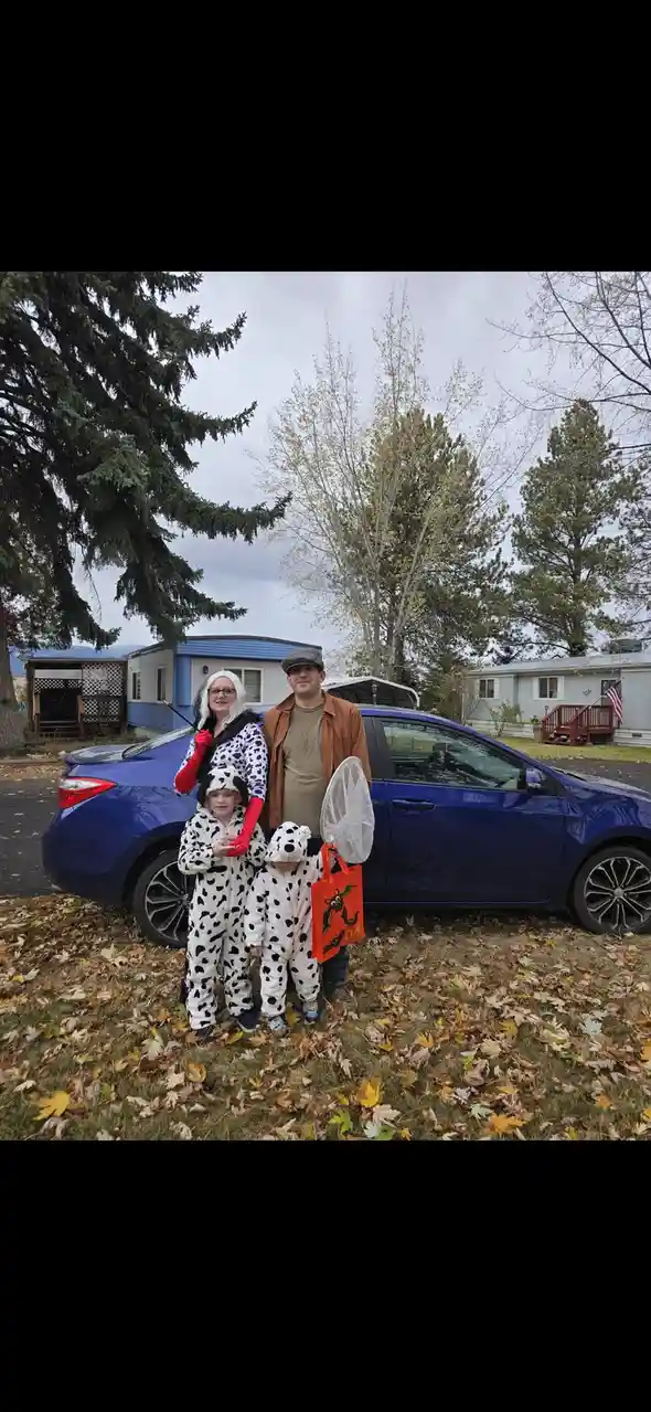 A family of four poses in front of a blue car in a yard with fallen leaves. The woman wears a Cruella de Vil costume with…