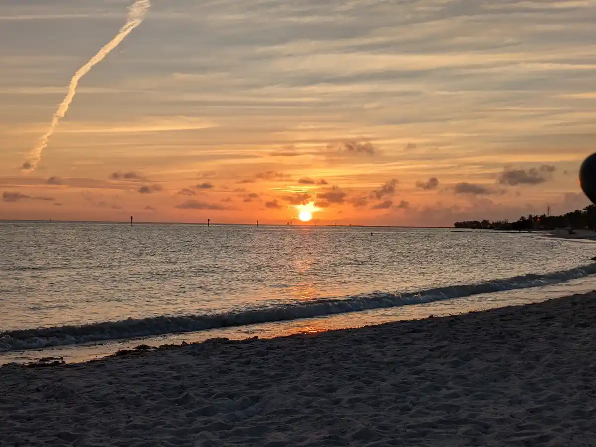 A serene beach sunset with orange and yellow hues reflecting on the water, a long contrail streaking across the sky, and a…