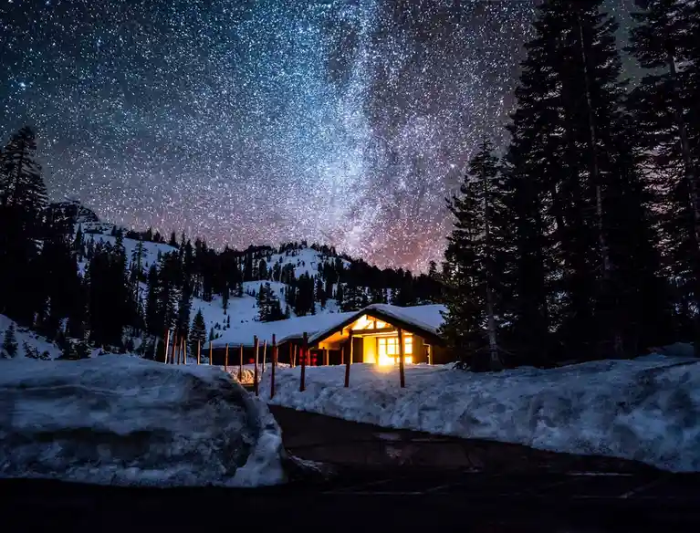A serene night scene shows a cozy cabin with warm lights glowing from its windows, nestled in a snowy forest under a…