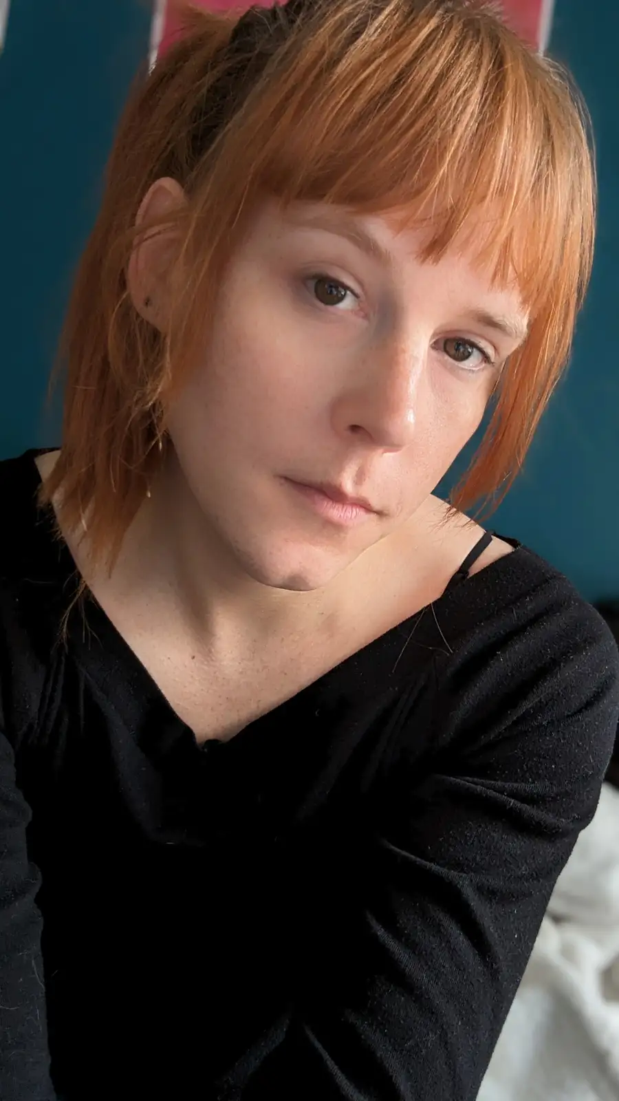 A person with reddish-brown hair and a black top stares directly at the camera with a neutral, slightly weary expression…