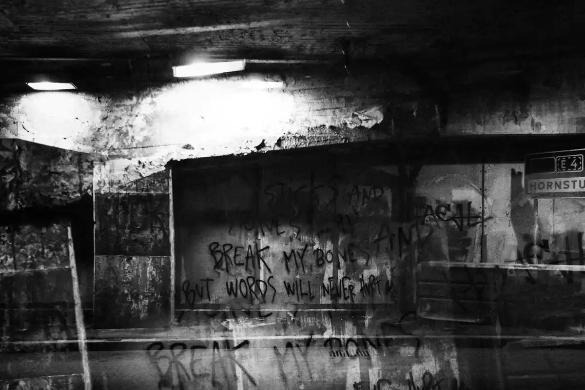 A black-and-white photo of a grimy, graffiti-covered subway wall under flickering fluorescent lights, with the text 'sticks…