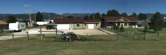 a wide shot of a modest single-story house with a brown roof and beige siding, set in a rural landscape with mountains in…