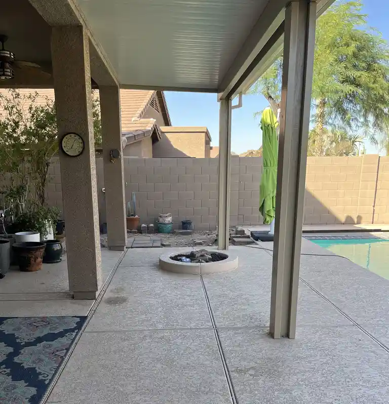 A backyard patio with a pool, fire pit, and a thermometer on a pillar showing 100 degrees. A green umbrella is closed, and…