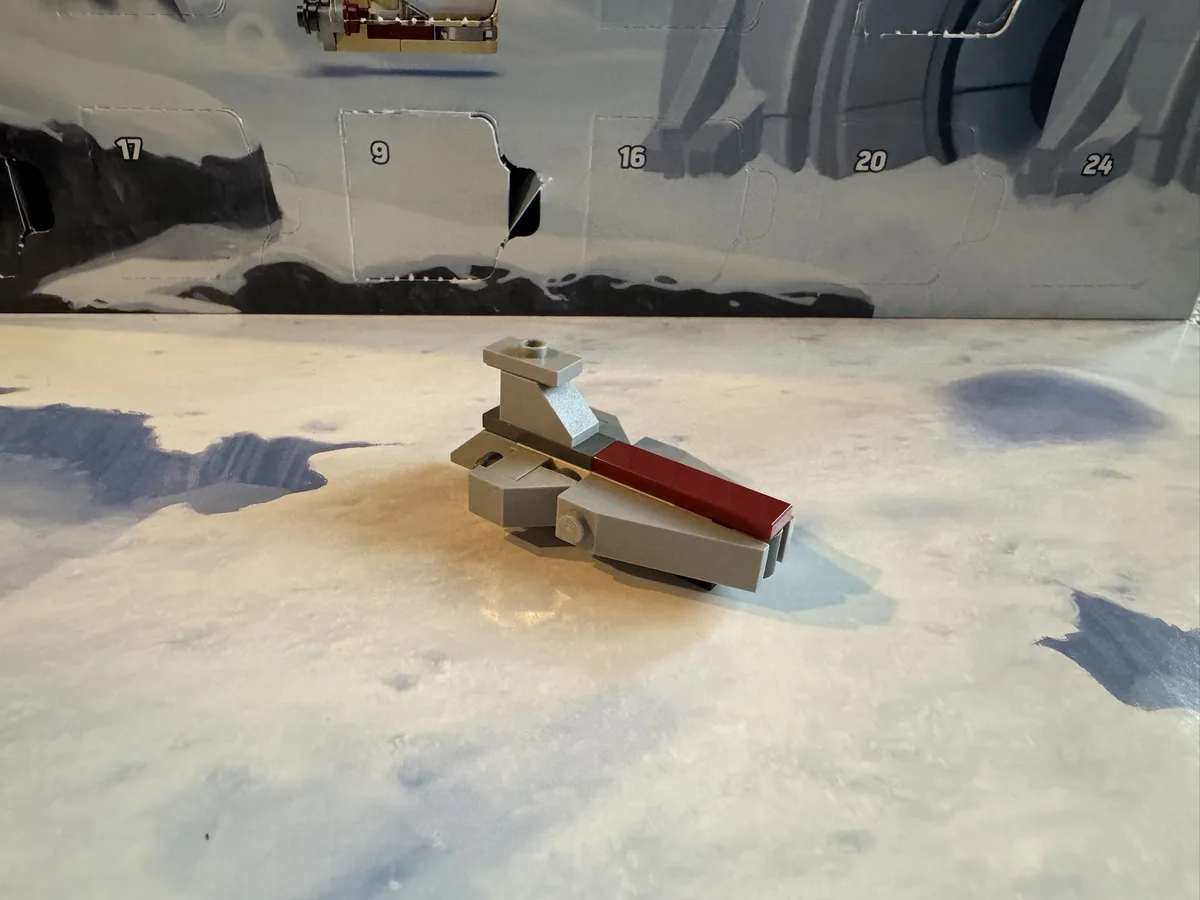 A small, gray Lego spaceship model with a red stripe sits on a surface printed with a snowy, mountainous landscape. Behind…