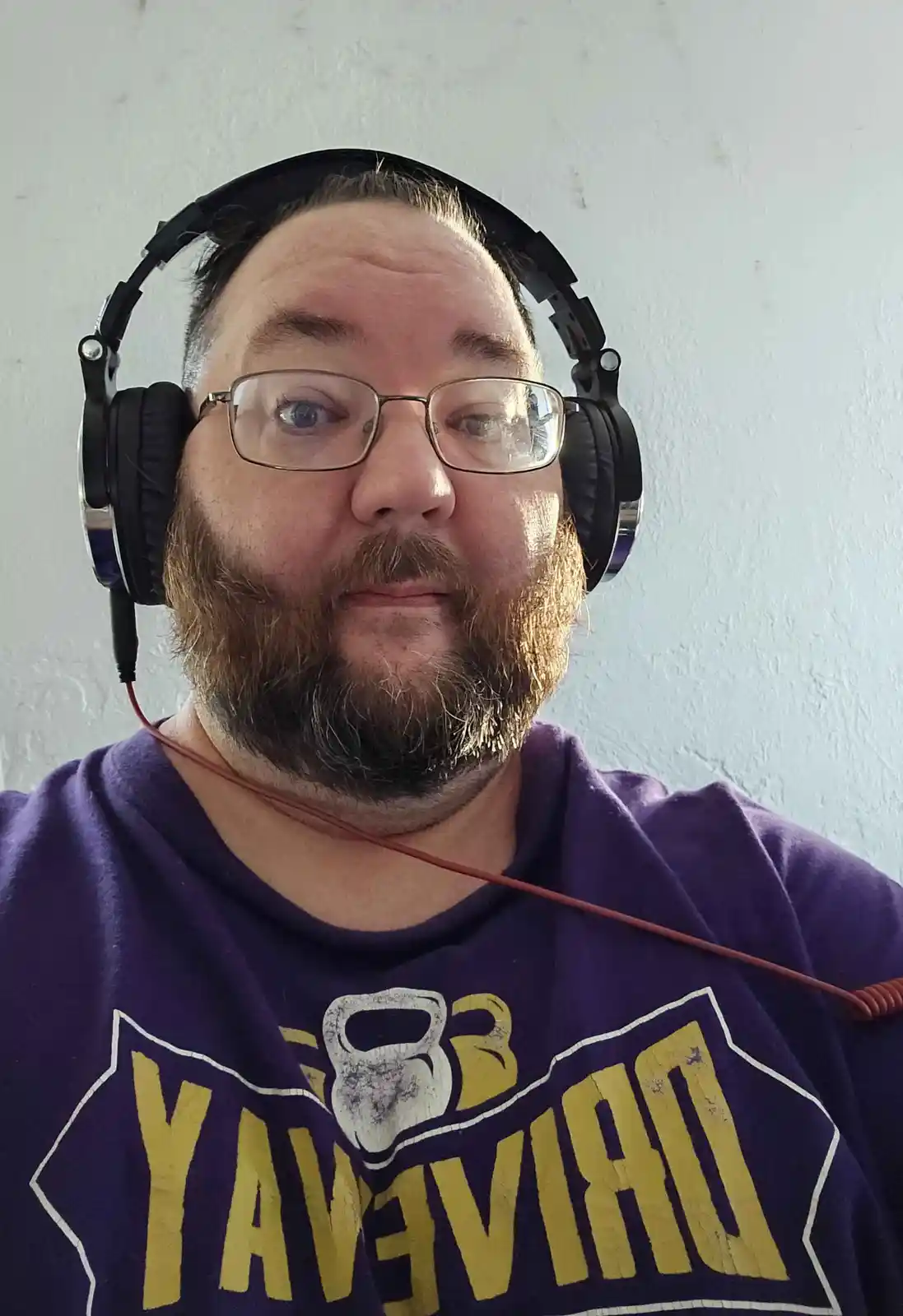 A man with glasses and a beard wears large black over-ear headphones while looking directly at the camera with a slightly…