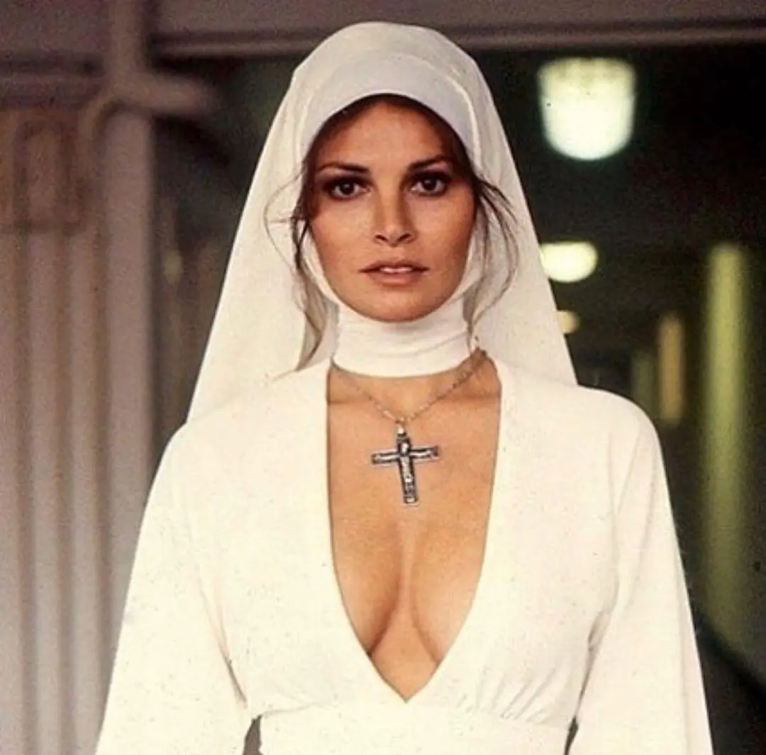 a woman in a white nun's habit with a deep V-neckline and a large cross necklace looks directly at the camera. The overlaid…