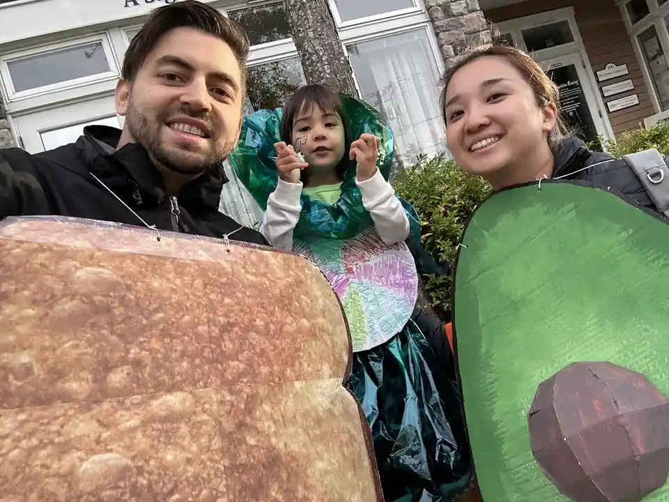 A family of three poses for a selfie outdoors, dressed in homemade Halloween costumes. The man on the left wears a large…