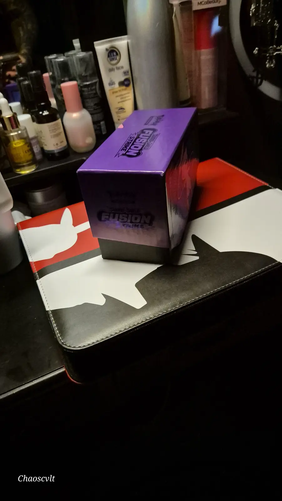 A purple Pokémon TCG 'Sword & Shield Fusion Strike' booster box sits atop a black, red, and white card sleeve featuring a…