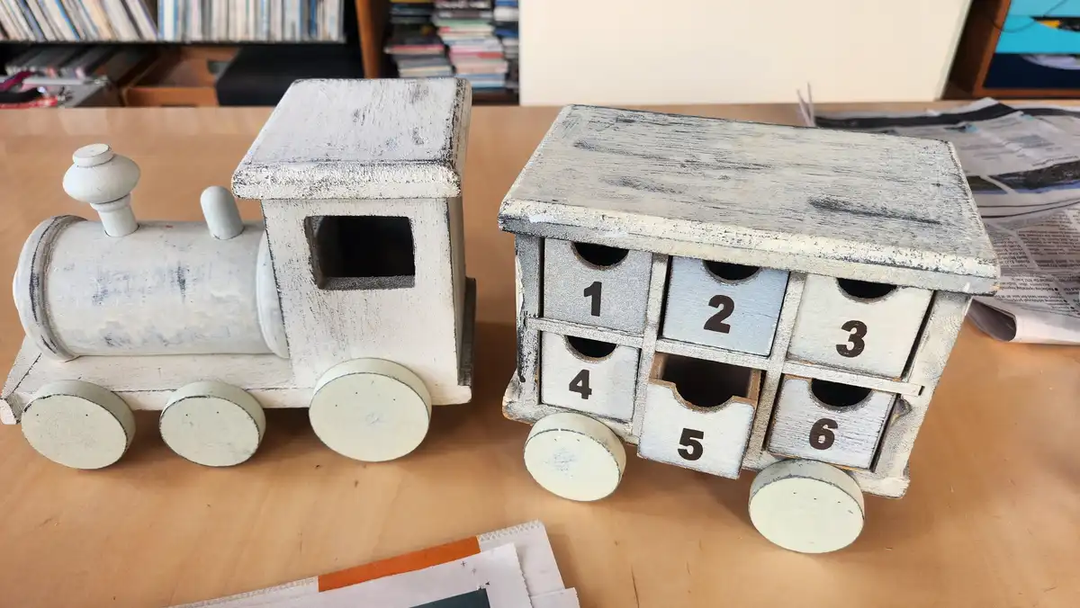 A white-painted wooden toy train with a locomotive and a numbered car sits on a wooden table. The car has six numbered…