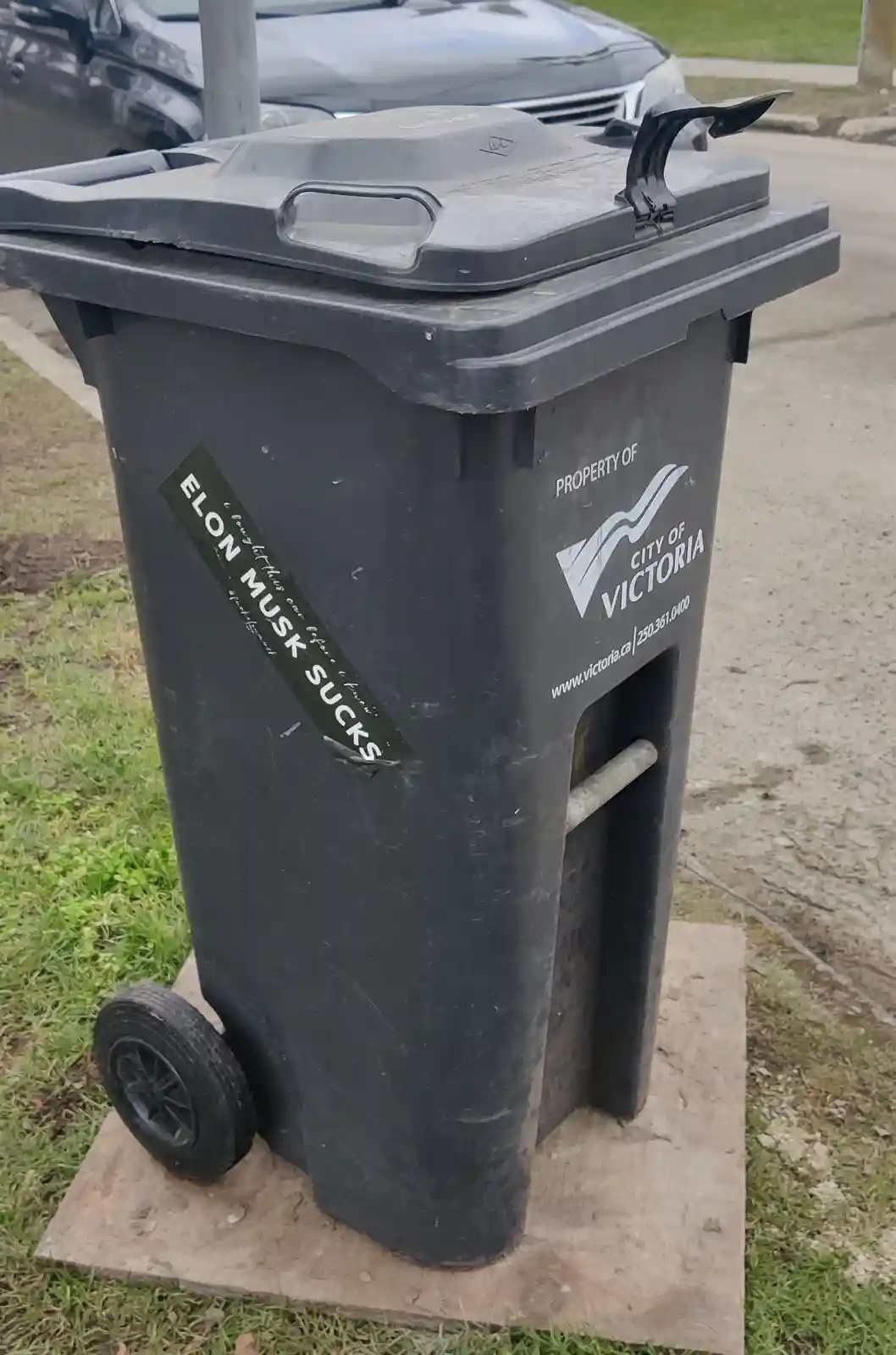 A black public trash can, labeled 'PROPERTY OF CITY OF VICTORIA', has a green sticker slapped on its side that reads 'ELON…