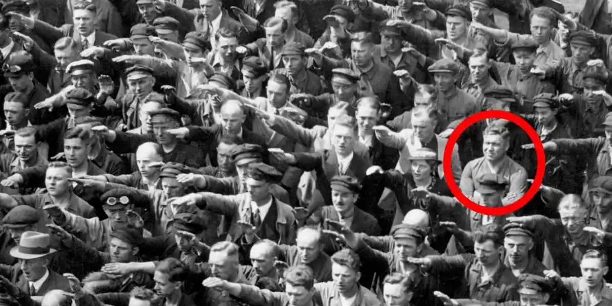 A black-and-white historical photograph shows a large crowd of men, most performing the Nazi salute with their right arms…